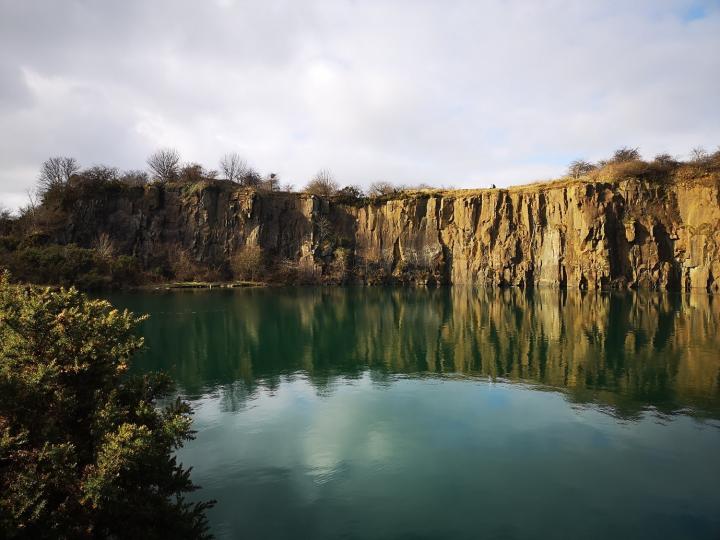 Photograph of a quarry, in the foreground is a calm pool of water. Behind is a large wall of rock with trees and bushes growing 