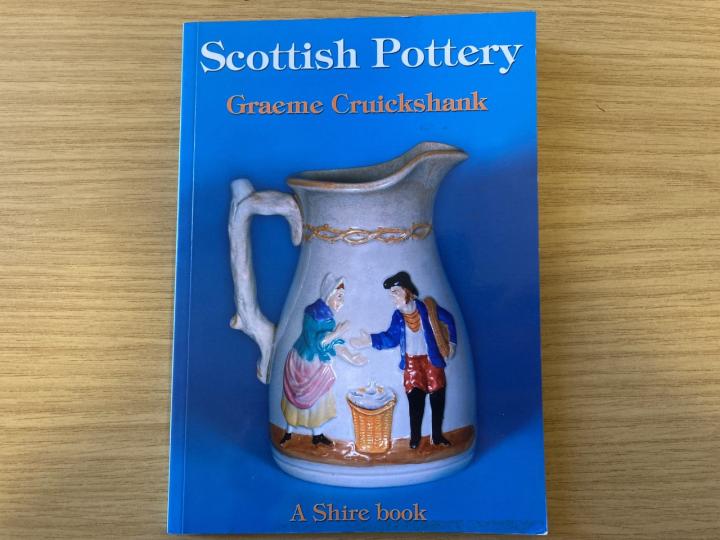 A photo of a book titled "Scottish Pottery" the front cover of the book shows a pot with two people enamelled on it.