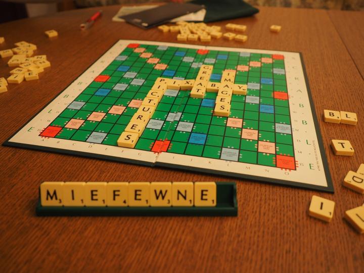 Photograph of a scrabble board on a brown table. Beside the scrabble board are different letter scrabble tiles. 