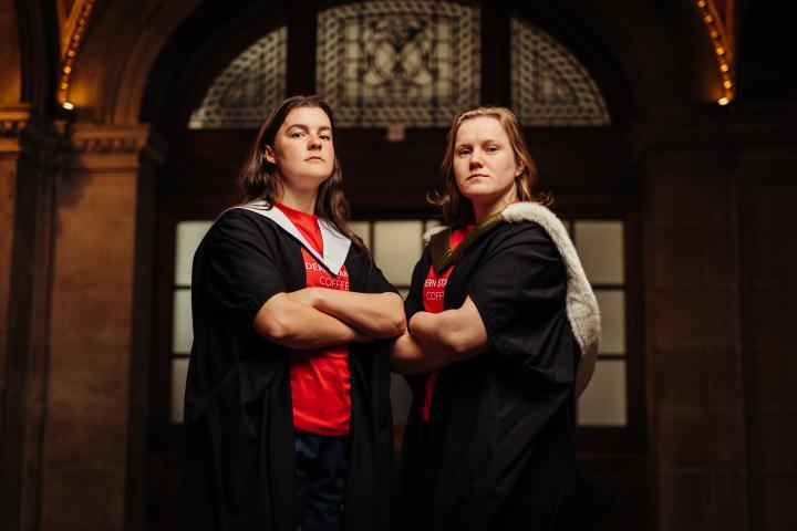 Sarah Denholm and Meryl Smith wearing graduate gowns and sport kit with arms crossed in McEwan Hall by stained glass windows