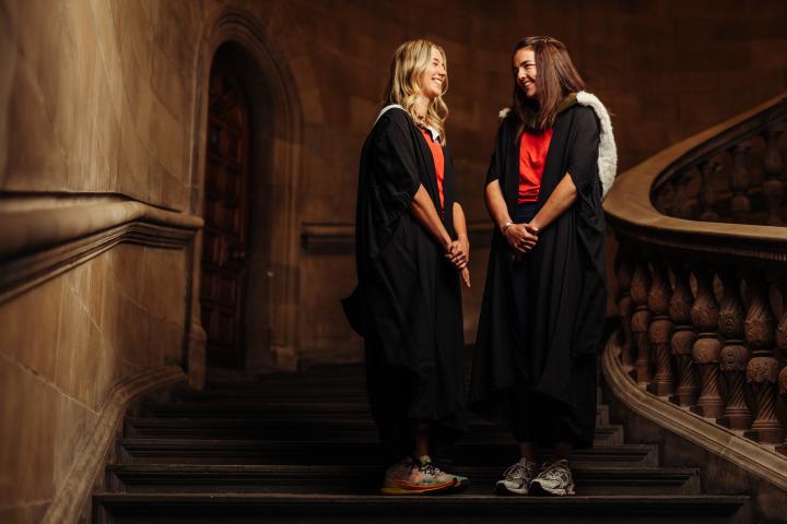 Eve and Claire wearing graduate gown and sports kit on McEwan Hall staircase