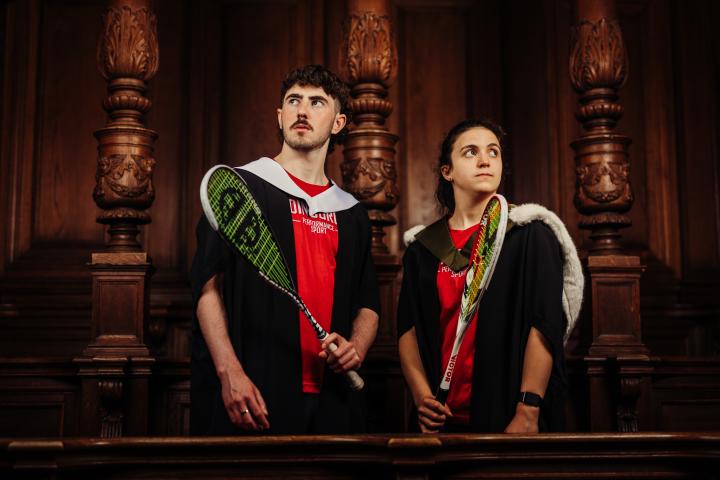 John Meehan and Georgia Adderley wearing graduate gowns and sports kit holding squash rackets on McEwan Hall stage