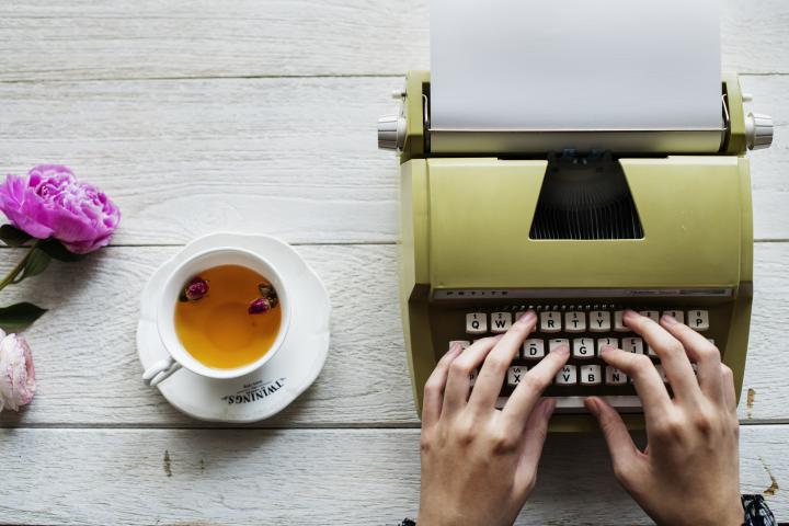 Someone typing on a typewriter, with a cup of tea beside them and some flowers. 