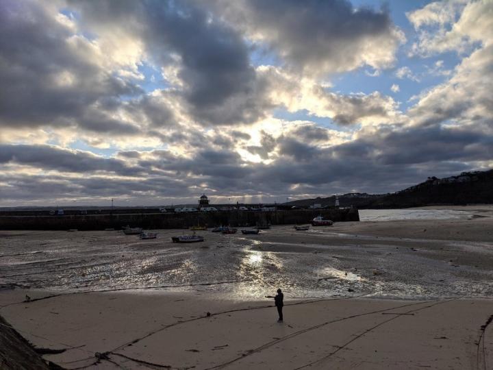 Sun breaking through the clouds at St Ives Harbour. In the foreground there is a person standing on a sandy beach, their is a harbour wall in the distance with the sea beyond that. Overhead their are light blue and grey clouds and sunlight coming through between them. 