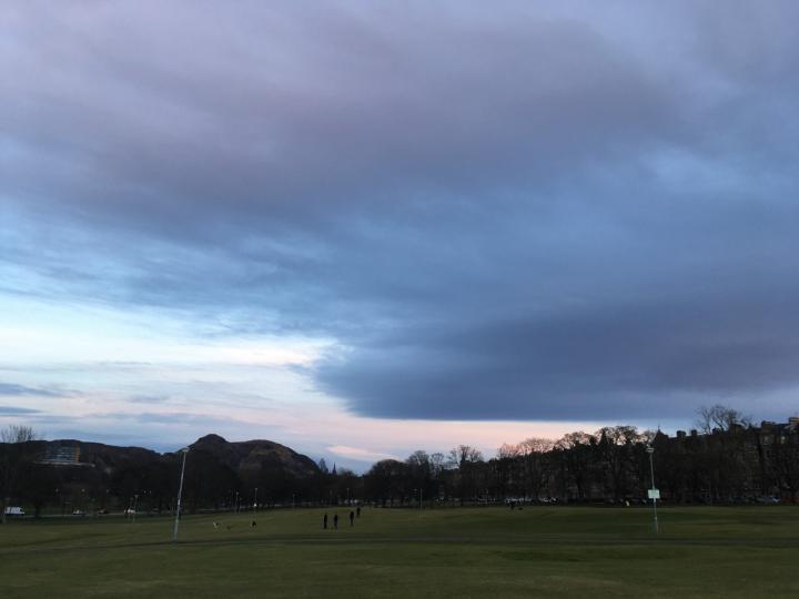 Photograph of The Meadows with Arthur's Seat in the background. Dark clouds meet lighter sky overhead.