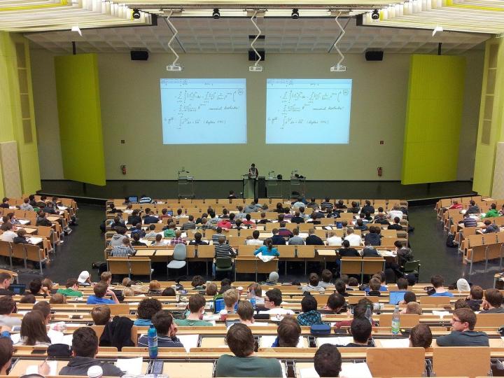 Photograph of a lecture theatre. They are students in seats listening to a lecture and a powerpoint on two screens projected onto a green wall