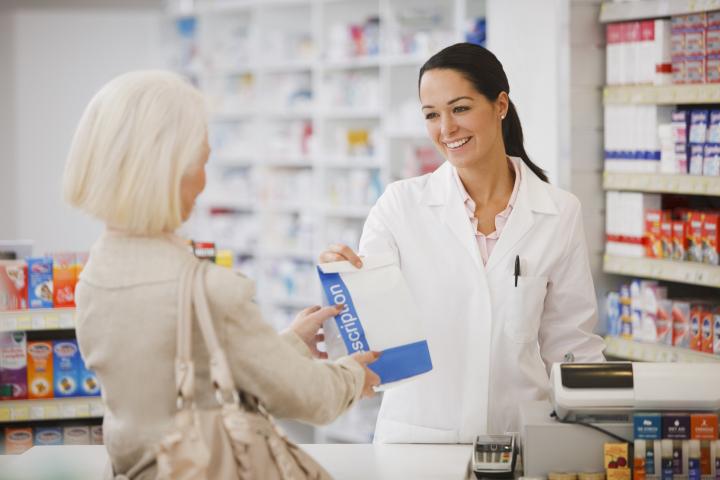 A female pharmacist with black hair and a white coat is handing over a prescription to a woman with a blond hair