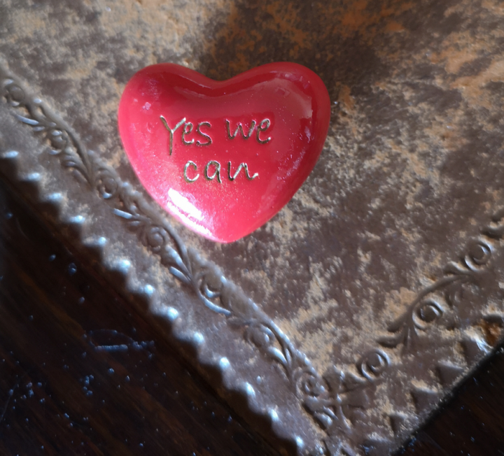 Photograph of a red stone in the shape of a heart with the words "Yes We Can" carved onto it. The red stone is sitting on the edge of a brown table. 