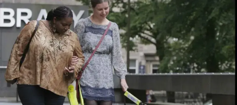 Blind student with guide dog and helper outside the University library