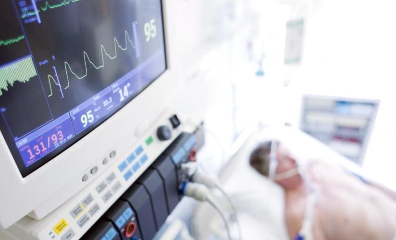 Close up of machines monitoring heart rate with man in bed in intensive care in background out of focus