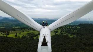 Two people in yellow hard hats stand on top a wind turbine and look at green countryside