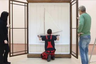 Two people watch the instalation of a model boat against white wooden paneling inside a dark wooden case with glass windows in the doors.