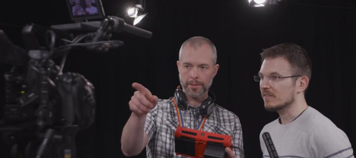 Two men are seen standing and talking together behind a camera