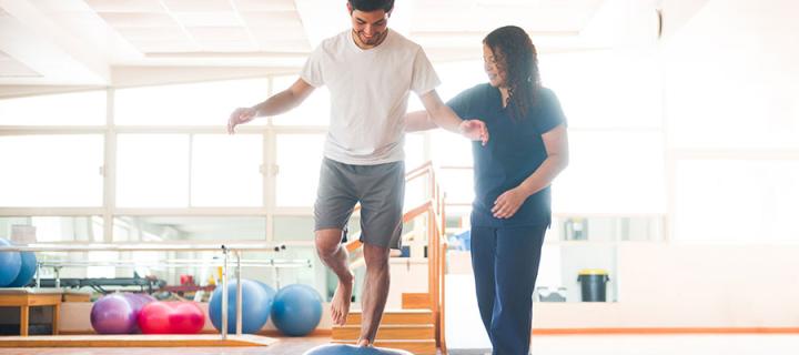 Young man standing on an balance trainer with one leg next to a female occupational therapist in a physical therapy room