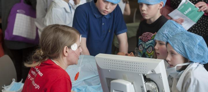 A group of children with wearing clinical protective clothes listening to an adult with electrical probes on their head talk