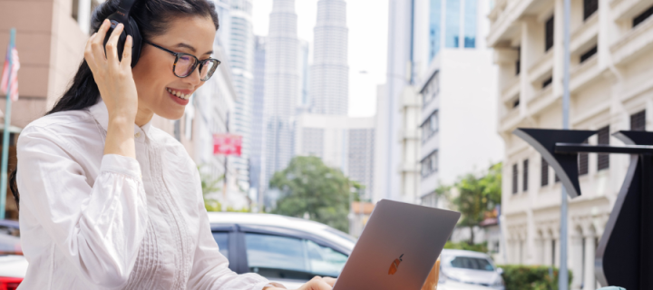Image of woman wearing headphones looking at her laptop in and outside city setting 