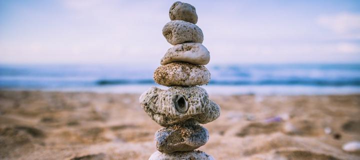 A photograph of stones stacked on top of each other on a beach. In the distance you can see the sea and blue sky overhead.