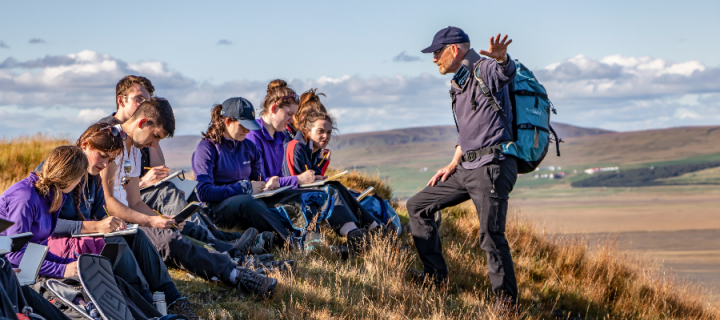 A male academic stands on a hillside and gestures to a group of students taking notes surrounded by the Icelandic landscape