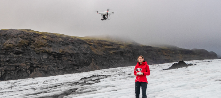 A female scientist in snow gear on a glacier in Iceland holding controls for a drone flying above her head
