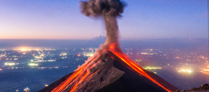 At night, an erupting volcano with smoke and lava in the foreground, with the lights of a major city of Guatemala behind
