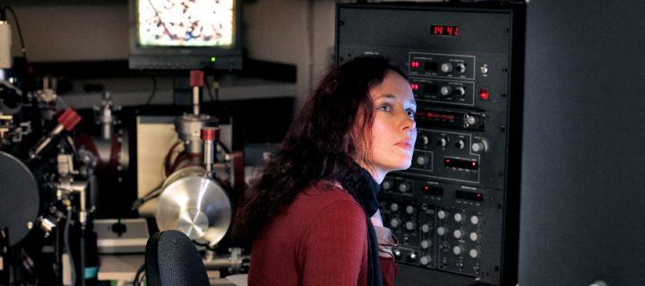 A female scientist seated at a desk surrounded by technical equipment in a science facility