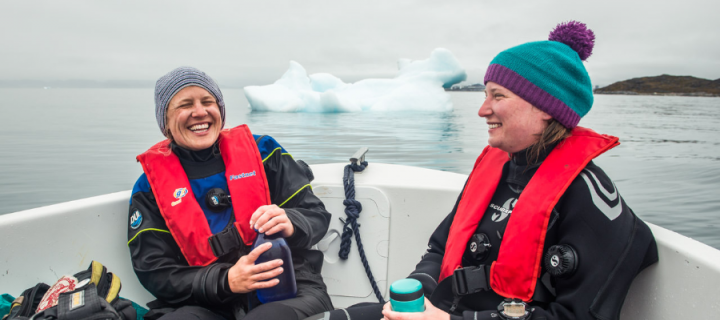 Two female scientists sitting in a small boat wearing cold water wetsuits with the ocean and an iceberg just behind them