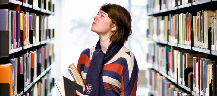 A female research student standing between two university library bookshelves, holding a book and looking at books