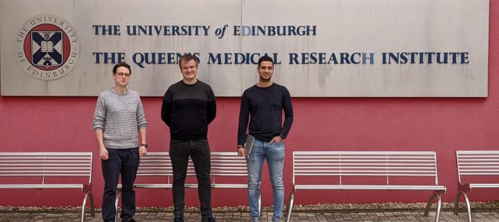 Three people standing outside a building, next to a sign that reads Queen's Medical Research Institute