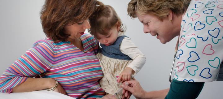 A girl sits in a woman's lap while having a vaccine jab in her left leg.