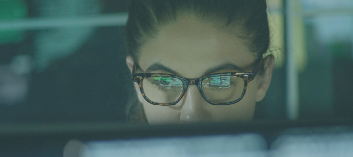 Image shows the top half of a woman wearing glasses looking at her laptop, rest of her face covered by laptop