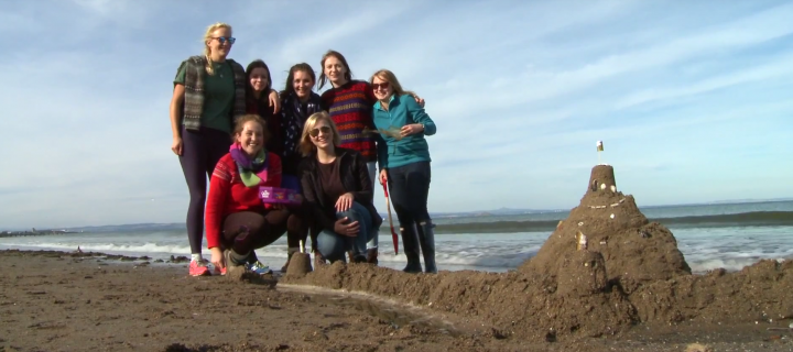 students stand next to a sandcastle they have built on a beach