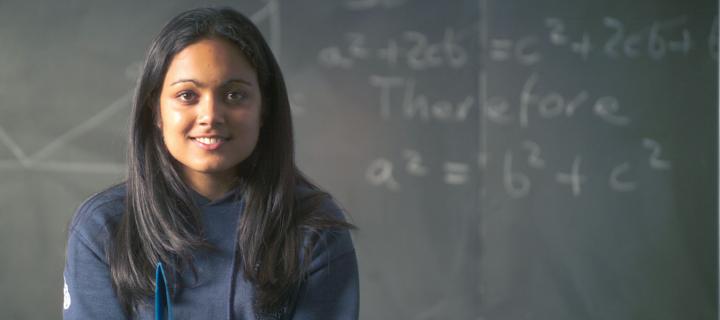 A girl sits in front of a blackboard with several equations on