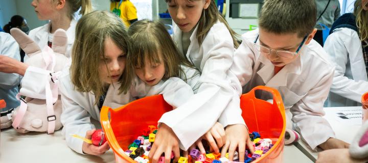 Several children wearing lab coats, gathered around an orange tub full of puzzle pieces