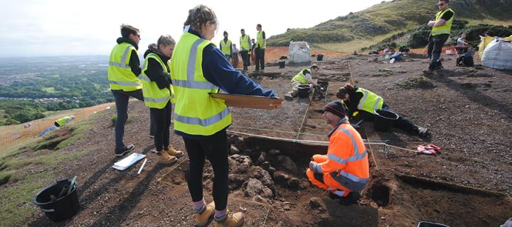 Students and professional archaeologists wearing high-vis clothing excavate and record a trench in Holyrood Park, Edinburgh