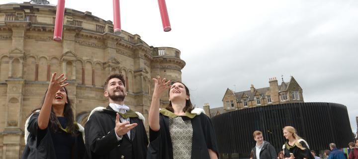 Three graduates standing together and celebrating their graduation by throwing red cylinder tubes in the air 