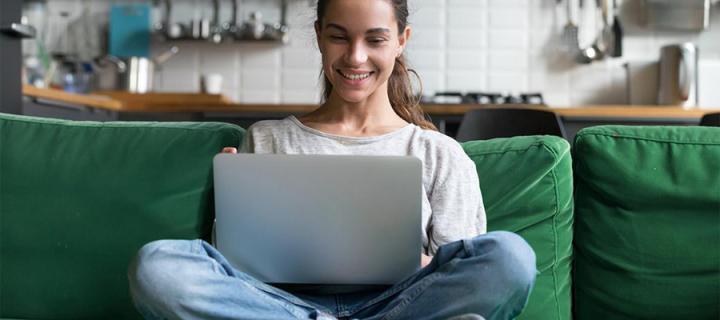 A woman sits cross legged on her couch smiling, working on her laptop that's on her lap