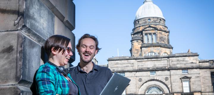 Two students with laptop chatting and smiling in front of Old College