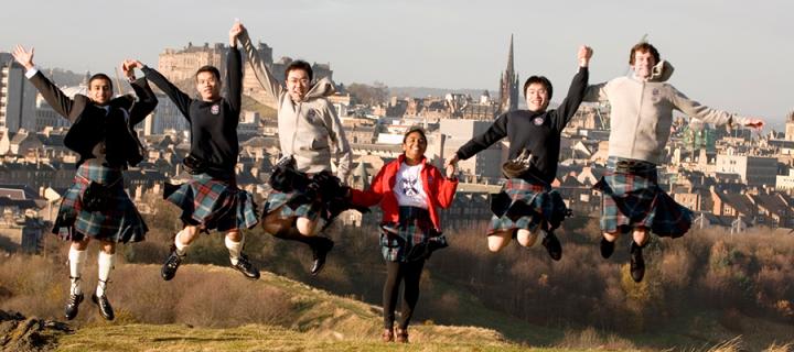 Group of students jumping outside, with a backdrop of the University campus.