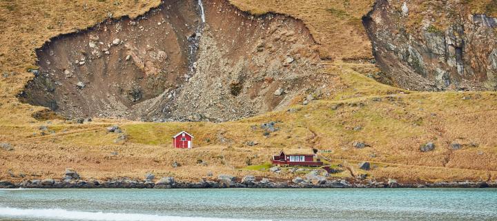 A red wooden barn sits at the base of a hill beside an icy blue lake. In the background, a large landslide has occurred.