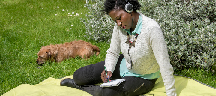 person sitting on blanket atop grass writing in notebook and listening to music with a dog laying nearby