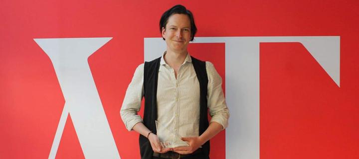 Photo of Patrick Errington holding his Emerging Writers award in front of a red and white screen