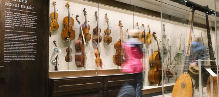 A blurred figure in a pink top walks past a museum display case containing string instruments