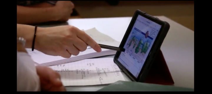 A hand points to a tablet, which some students are learning from.