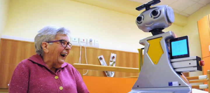 Image shows an elderly woman sat down, laughing besides a robot.