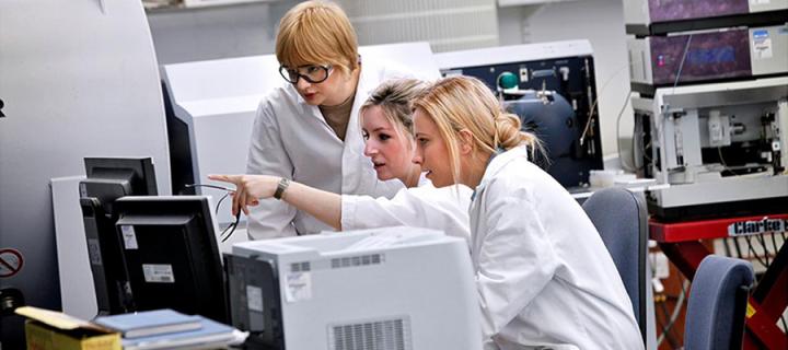 Three people in lab, wearing lab coats study images on a screen