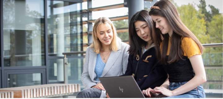 image of the female students look at a laptop sitting on a bench in an outdoor setting