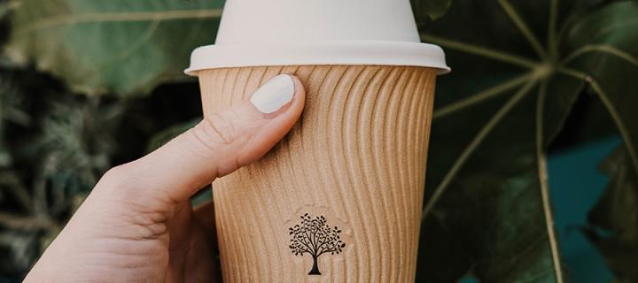 Hand holding a cardboard coffee cup with a tree picture on it, against a background of large leaves