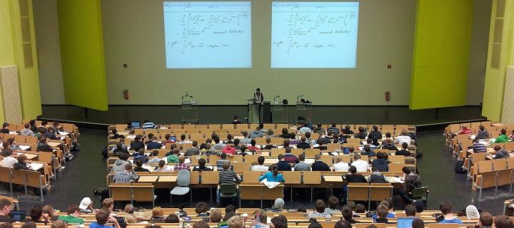 Photograph of a lecture theatre. They are students in seats listening to a lecture and a powerpoint on two screens projected ont