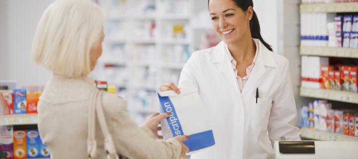 A female pharmacist with black hair and a white coat is handing over a prescription to a woman with a blond hair