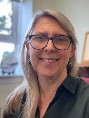 Head and shoulders of a smiling woman with glasses. Light coloured hair is over her right shoulder and she is wearing a dark shirt.